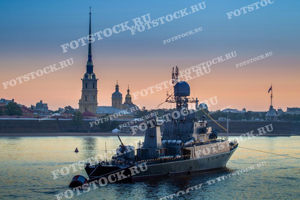 На фотографии изображён военный корабль на фоне рассвета в Санкт-Петербурге. Вдалеке виднеется шпиль Петропавловской крепости, окутанный мягким светом утренней зари. Небо переливается оттенками розового и голубого, что создаёт спокойную и величественную атмосферу. Освещённые огни корабля добавляют картине живости и детализации. Эта фотография может использоваться в изданиях, посвящённых военно-морской тематике, туристических путеводителях, в качестве учебного материала по истории или для украшения интерьера.